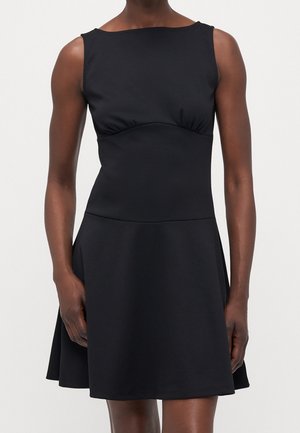 Czarna sukienka bez rękawów z dopasowaną górą i rozkloszowanym dołem, noszona przez stojącą osobę na jednolitym tle.