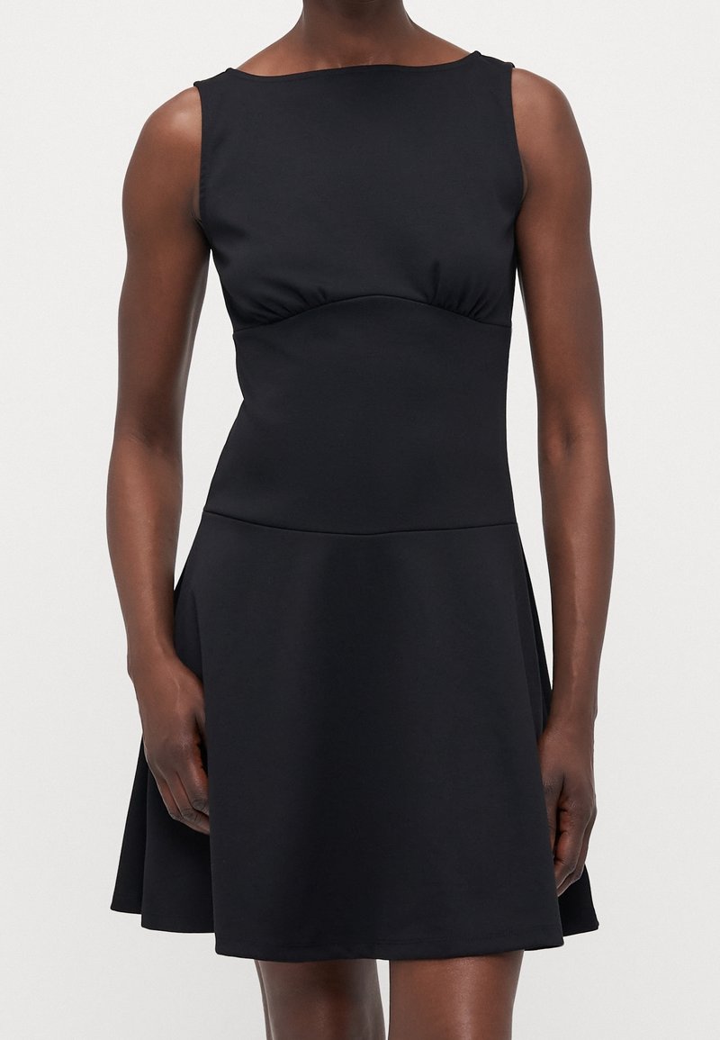 Robe noire sans manches avec un corsage ajusté et une jupe évasée portée par une personne debout devant un fond uni.