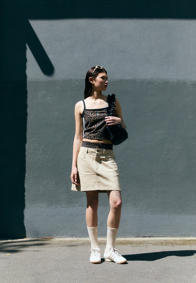 Top court à motifs léopard avec des bretelles noires, jupe en denim beige, sac à main noir, chaussettes blanches et baskets claires, se tenant devant un mur gris.