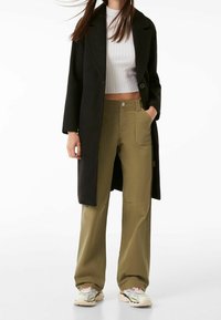 Černý dlouhý kabát přes bílý žebrovaný krátký top, doplněný olivově zelenými širokými kalhotami a v ícebarevnými teniskami, které ukazují kontrast textur.