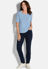 Glunderende vrouw die staat met één hand in haar zak, gekleed in een lichtblauwe, gestructureerde top met korte mouwen, donkere jeans en witte sneakers op een witte achtergrond.