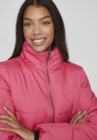 Jachetă puffer roz cu guler înalt, fermoar frontal și detalii negre. Materialul pare neted, cu un design cusut. Are o croială relaxată.