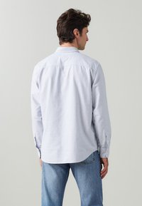 Chemise à rayures boutonnière en bleu clair et blanc, fabriquée dans un tissu à texture douce. Elle possède un col et des poignets boutonnés, avec une coupe ajustée.