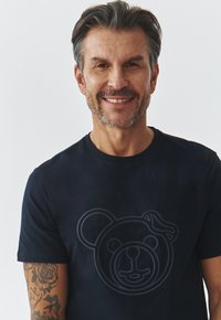 Juodas medvilnės T-shirt su tamsiu meškos grafiku priekyje. Trumpi rankoviai ir apvalios kaklo dizainas. Lygus paviršius ir laisvas siluetas.