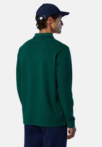 Polo de manga larga verde con cuello clásico, textura suave y dobladillo recto. Combinado con gorra y pantalones de color azul marino oscuro.