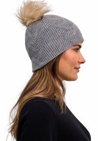 Vrouw in zwarte trui met een grijze gebreide muts met pompon, gezien vanaf de rechterzijde op een witte achtergrond.