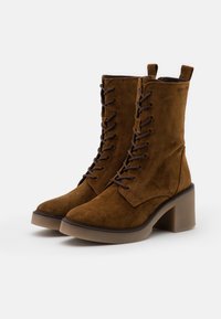 Brązowe zamszowe botki za kostkę z wiązaniem z przodu, zaokrąglonym noskiem i masywnym przezroczystym obcasem. Charakteryzują się przeszywanym wzorem i pętelką do ściągania.
