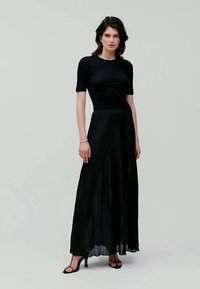 Vestido maxi preto plissado com um corpete justo de manga curta e tecido translúcido. Combinado com sandálias de salto preto, em um fundo neutro.