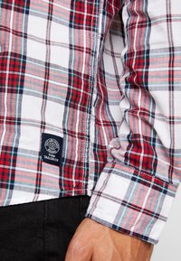 Ingrandimento di una manica di una camicia a quadri rossa, bianca e blu con un'etichetta scura del marchio Tom Tailor e una mano appoggiata accanto a dei pantaloni scuri.