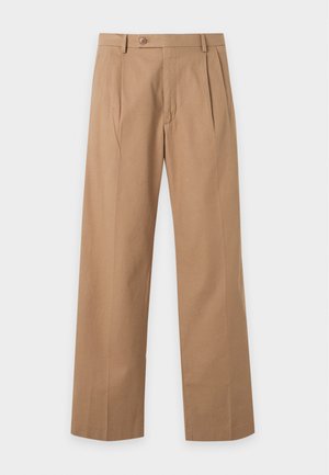 Jonge man staand, gekleed in een zwart lang mouw shirt, beige wijde broekspijpen en zwarte sneakers met witte zolen op een witte achtergrond.