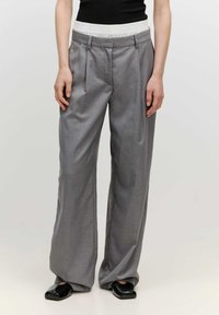 Graue weit geschnittene Hose mit einem strukturierten Bund, ausgestattet mit Vorderfalten und einem glatten Stofffinish. Kombiniert mit glänzenden schwarzen Schuhen.