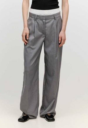Pantalons larges gris avec une taille texturée, dotés de plis sur le devant et d'une finition en tissu lisse. Associés à des chaussures noires brillantes.