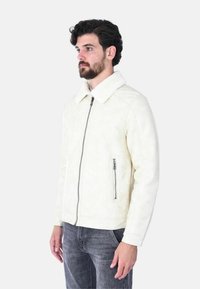 Giacca in shearling bianco con un colletto morbido, chiusura a zip, tasca laterale con zip e texture liscia. Il modello la indossa sopra jeans grigi.