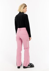 Roze skibroek met een gladde textuur, voorzien van ritssluitingen in de zakken en uitlopende pijpen, gecombineerd met een zwart geribd longsleeve shirt en skischoenen.