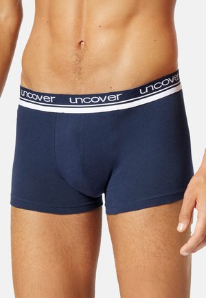 Close-up of a man’s torso and upper legs wearing navy blue boxer briefs with a white elastic waistband labeled "uncover."