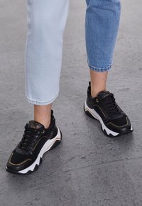 Sneaker nere con suole bianche e dettagli dorati; presenta una tomaia in sintetico liscio, punta tonda e sistema di allacciatura. Indossate con jeans azzurri chiari.