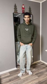 Φούτερ με κουκούλα σε ελιά πράσινο με το κείμενο "Barbour", ανοιχτό μπλε τζιν και λευκά αθλητικά παπούτσια. Καθρέφτης ολόσωμος αντανακλά την εμφάνιση. Ουδέτερο φόντο.