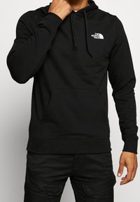 Mężczyzna w czarnej bluzie z kapturem The North Face i czarnych spodniach cargo, z jedną ręką przy twarzy, a drugą swobodnie opuszczoną wzdłuż ciała.