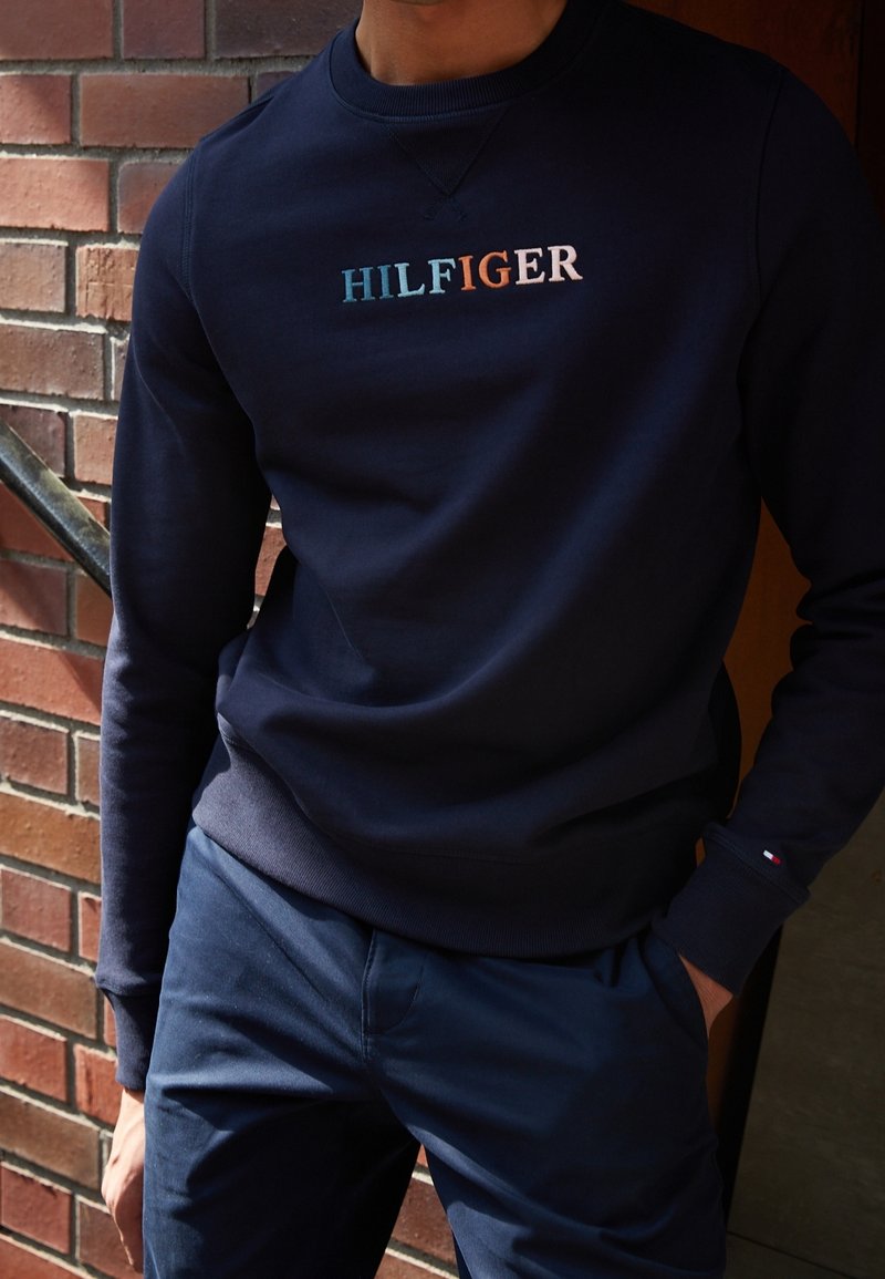 Marinblå sweatshirt med rund hals och långa ärmar. Har broderad "HILFIGER"-logga i ljusblått, orange och vitt. Mjuk tygstruktur.