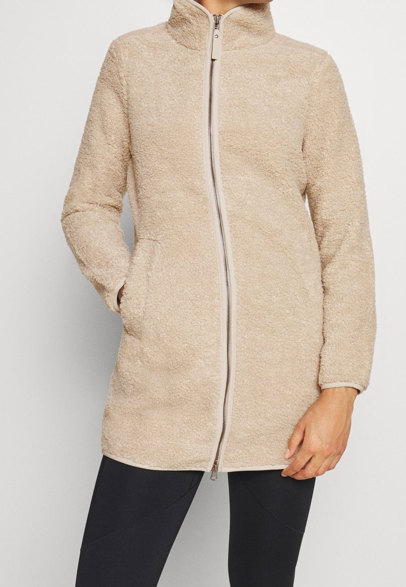 Beige fleece takki, jossa on korkea kaulus, täysipitkä vetoketju ja kaksi sivutaskua. Ominaisuuksiin kuuluu pehmeä rakenne ja istuvat hihat.