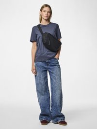 Kobieta w granatowej koszulce i szerokich niebieskich dżinsach stoi z jedną ręką w kieszeni, nosząc czarną torbę na ramię i bordowe buty.