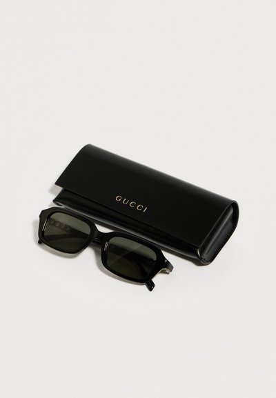 Lunettes de soleil rectangulaires noires avec verres foncés posées à côté d'un étui Gucci noir avec logo doré sur fond blanc.