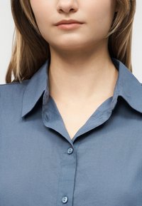 Νέος με ανοιχτόχρωμο δέρμα και καστανόξανθα μαλλιά μέχρι τον ώμο, ντυμένος με μπλε πουκάμισο με κουμπιά, κομμένο στο λαιμό και το στήθος.