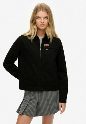 Veste zippée noire avec une poche avant et un patch logo, associée à une jupe grise à rayures. Le matériau semble lisse et léger.