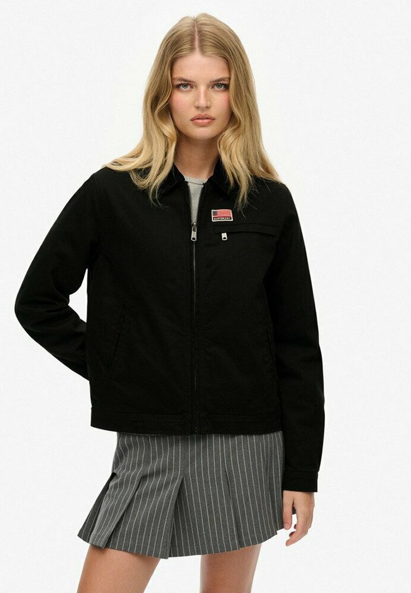 Sort zip-up jakke med en lomme foran og logo-patch, parret med en grå stribet nederdel. Materialet ser glat og let ud.