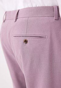 Jasnofioletowe spodnie z teksturowanej tkaniny, z tylną kieszenią zapinaną na brązowy guzik i szlufkami na pasek, widoczne z boku.