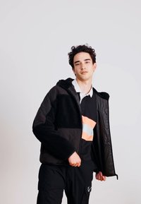 Veste noire avec un devant texturé et duveteux, des manches lisses et une capuche ; portée sur un t-shirt noir avec une bande orange et blanche.