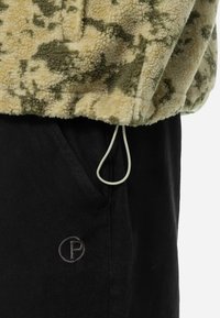 Fleece pullover med et beige og grønt camouflagemønster, udstyret med en elastisk snor, samt sorte sweatpants med et logo på lommen.