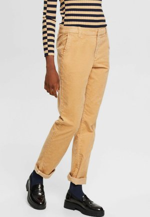 Person trägt beiger Cordhose, die an den Bündchen umgeschlagen sind, ein langärmliges, schwarz-beige gestreiftes Oberteil, schwarze Loafer und dunkle Socken.