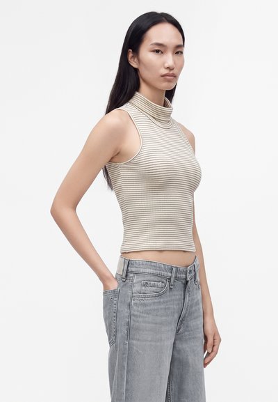 Jeune femme aux longs cheveux noirs portant un col roulé sans manches beige rayé et un jean gris délavé taille haute, une main dans la poche.