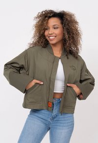 Olivgrüne Bomberjacke mit Reißverschluss, geripptem Kragen, Vordertaschen, eingesticktem Logo und gesteppter Textur; getragen über einem weißen Oberteil.