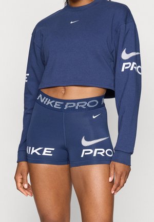 Frau trägt dunkelblaue Nike Pro Shorts und passendes gekürztes Langarmshirt mit weißen Nike-Logos und Texten auf Ärmeln, Shorts und Bund.