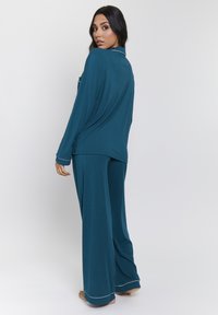 Teal pajama set featuring a loose long-sleeve top with a pocket and wide-leg pants, accented with light-colored trim. Soft textured fabric.