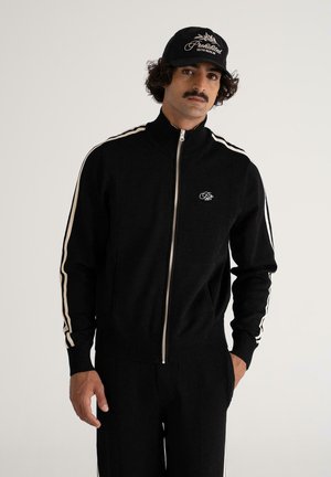 Man with mustache wearing black zip-up jacket with white stripes on sleeves, matching black pants, and black cap with white lettering.
