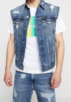 Mand iført distressed blå denimvest over hvid t-shirt med grafik og matchende revnede blå jeans, stående foran en hvid baggrund.