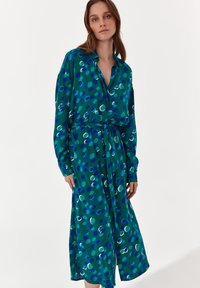 Abito verde con motivo a pois blu e bianchi, maniche lunghe, chiusura a bottoni frontale, vita cinte con una cintura, tessuto leggero, lunghezza media.