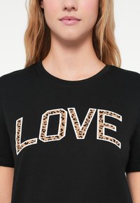 Μαύρο βαμβακερό Τ-σορτς με τη λέξη «LOVE» σε λευκό χρώμα, διακοσμημένη με καφέ λεοπάρ σχέδιο. Στρογγυλή λαιμόκοψη και κοντά μανίκια.