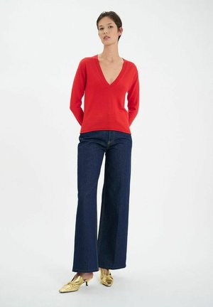 Pull en tricot rouge avec un col en V, associé à un pantalon en denim foncé à taille haute et à des mules à talons dorés. Fond uni.