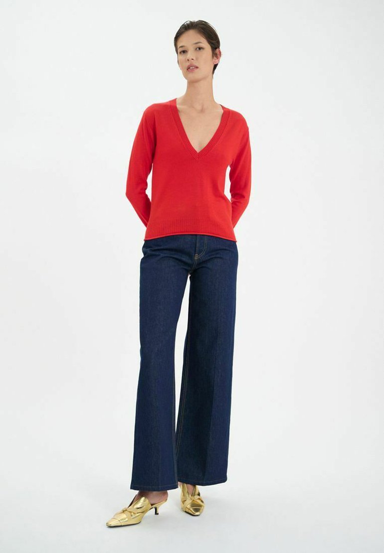 Pull en tricot rouge avec un col en V, associé à un pantalon en denim foncé à taille haute et à des mules à talons dorés. Fond uni.