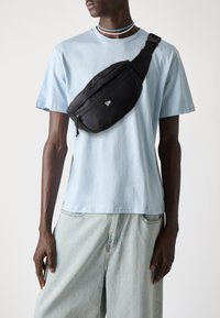 Sac bandoulière noir en matériau lisse, avec une poche zippée et un logo. Porté par-dessus un t-shirt bleu clair.