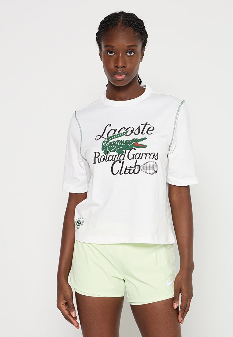 Jeune femme portant un t-shirt blanc Lacoste Roland Garros Club et un short Nike vert clair, se tenant contre un fond uni.