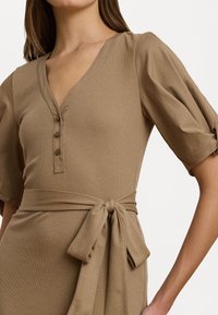 Abito in maglia color sabbia caratterizzato da scollo a V, dettagli con bottoni, maniche a palloncino e una cintura annodata in vita, evidenziando una texture morbida e un design semplice.