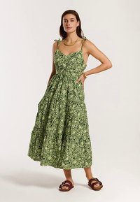 Zaļas ziedu raksta kleita ar regulējamām aukliņām, pieguļošu augšdaļu un pakāpienu svārkiem; izgatavota no viegla auduma ar dabīgu tekstūru.