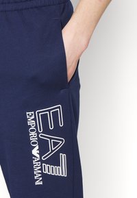 Marina blå sweatpants i mjukt tyg med sidofickor; har en vit tryckt logotyp "EA7 Emporio Armani" på vänster lår.