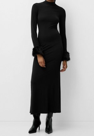 Vestido largo ajustado negro con cuello alto, mangas largas con puños de piel negra, combinado con botas negras de tacón alto y punta afilada.