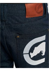 Dark denim jeans feature a thick texture and a prominent white graphic of an animal within a circular design on the back pocket. Brown leather label attached.